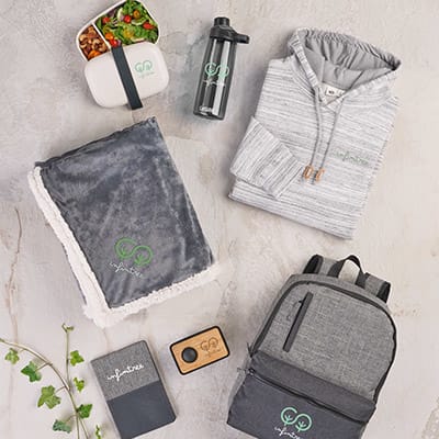 An image of A collection of branded items including a grey hoodie, fleece blanket, reusable water bottle, lunch container, notebook, bamboo Bluetooth speaker, and a grey backpack, all with eco-friendly branding displayed on a light surface.