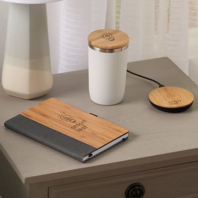 An image of desk setup featuring a bamboo and fabric notebook, a white insulated cup with a bamboo lid, and a bamboo wireless charging pad, all branded with an eco-friendly logo.